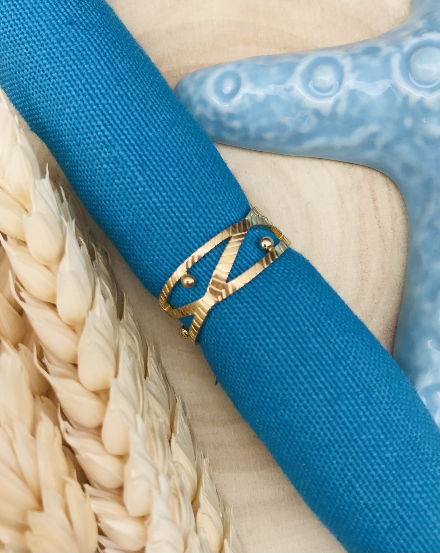 Guardanapo azul com anel dourado ao lado de espigas secas e estrela do mar azul clara.