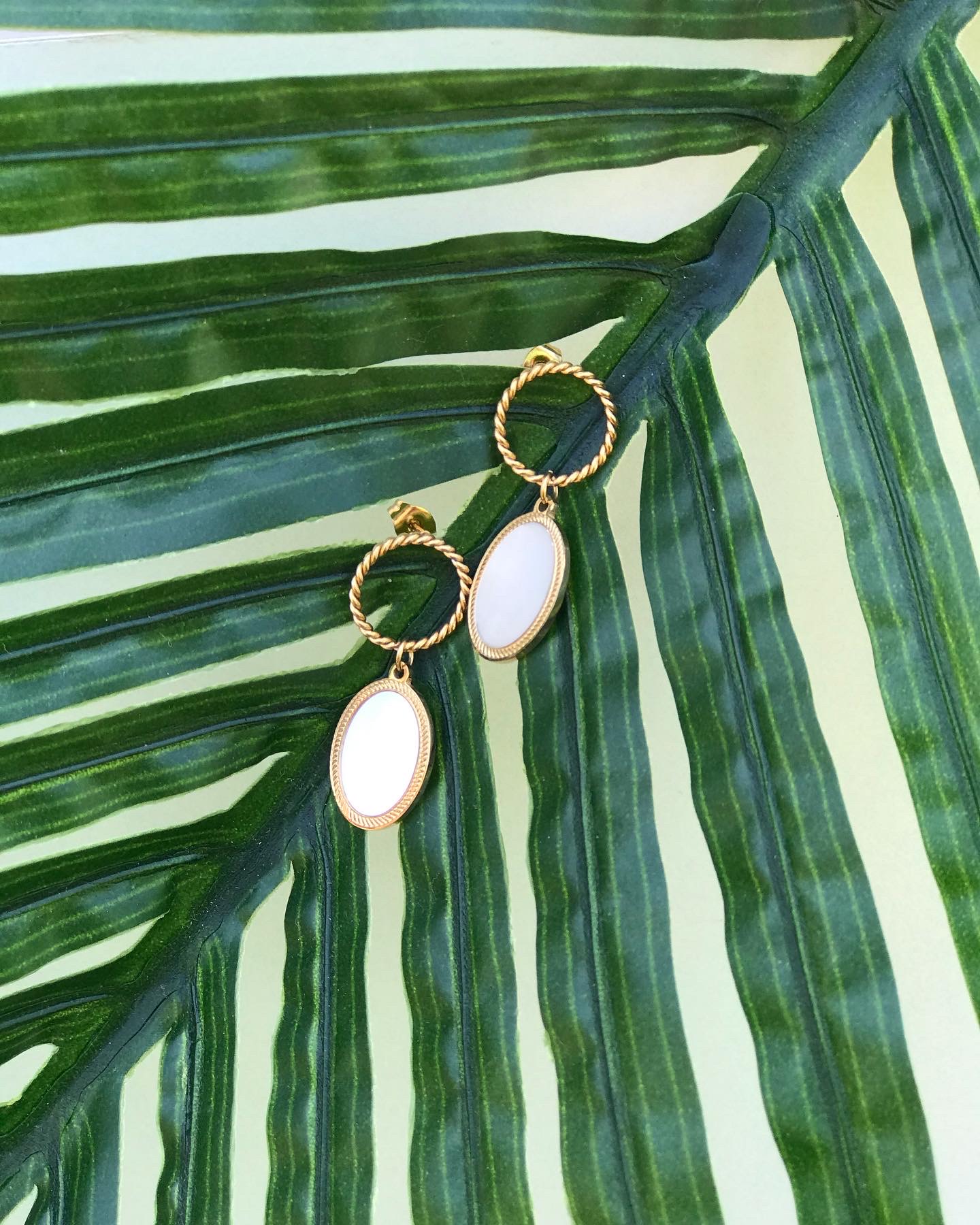 Brincos dourados com pendentes ovais brancos sobre folha verde
