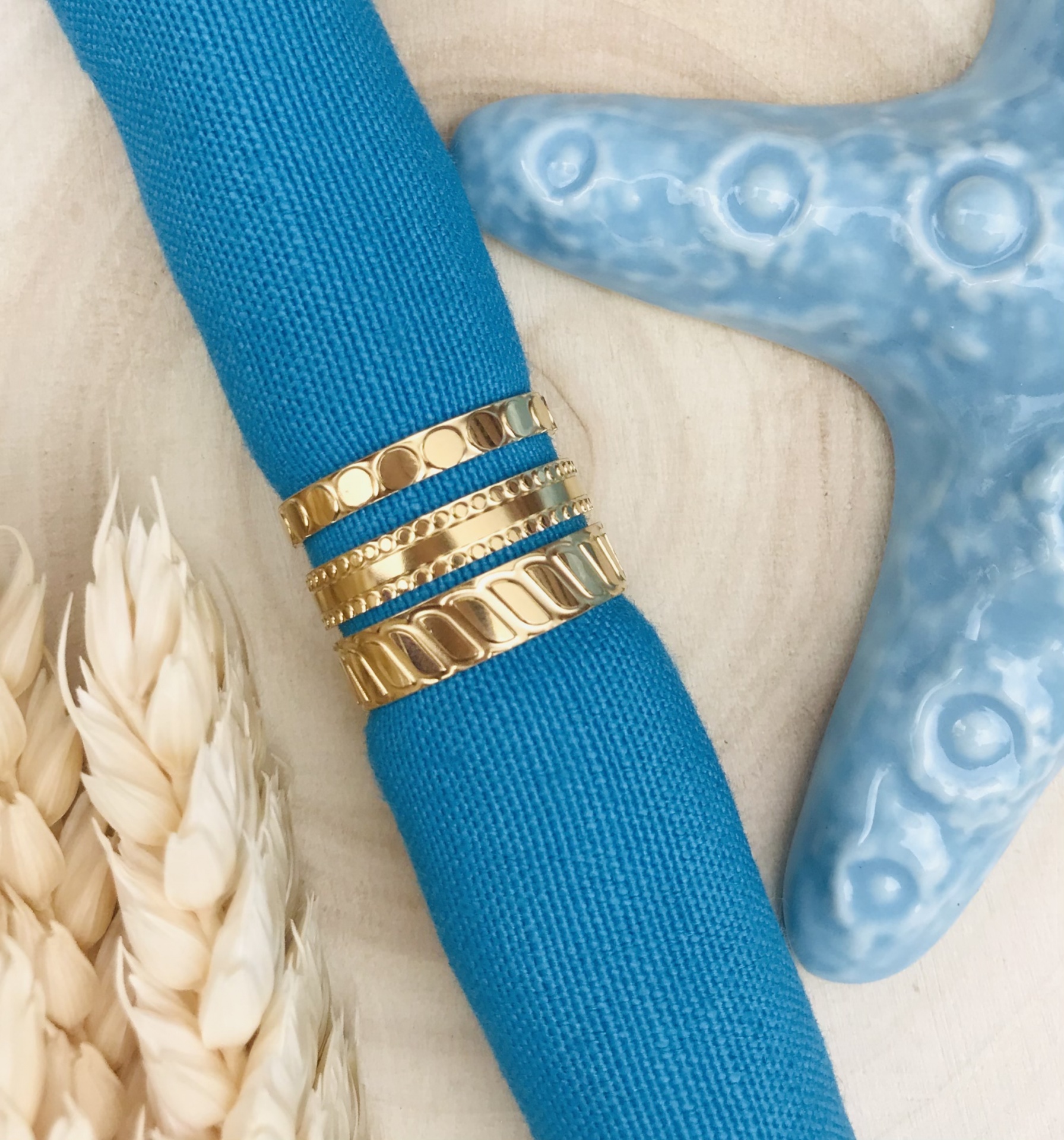 Cinco argolas douradas com padrões variados sobre pano azul, próximo de estrela-do-mar cerâmica e planta seca