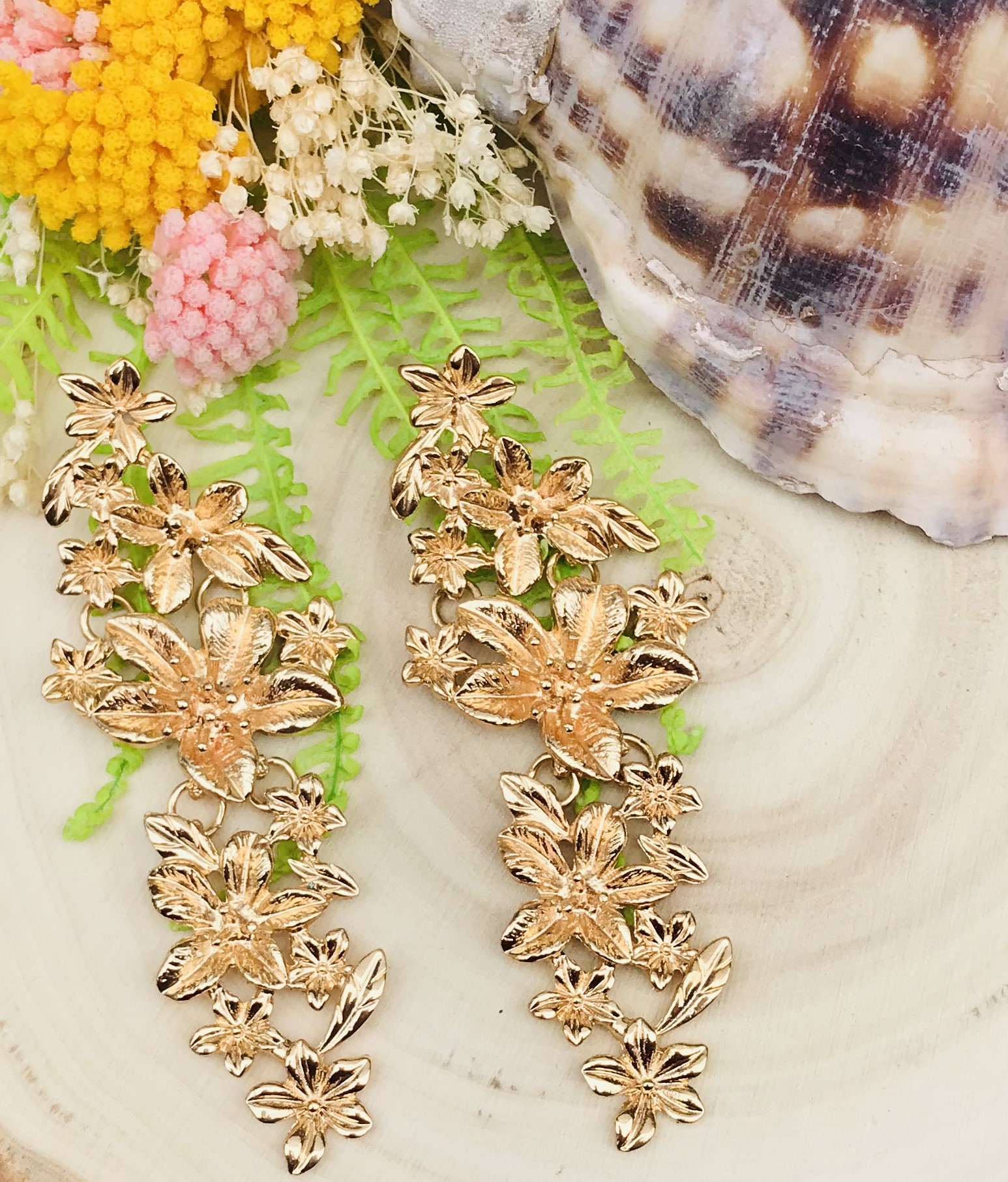 Brincos dourados longos com flores em fundo com plantas, flores secas e concha