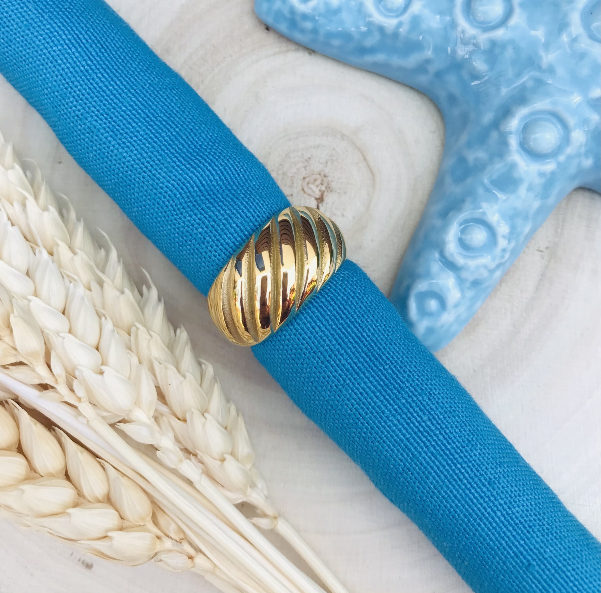 Pulseira azul em tecido com fecho dourado texturizado sobre madeira com estrela do mar azul e trigo