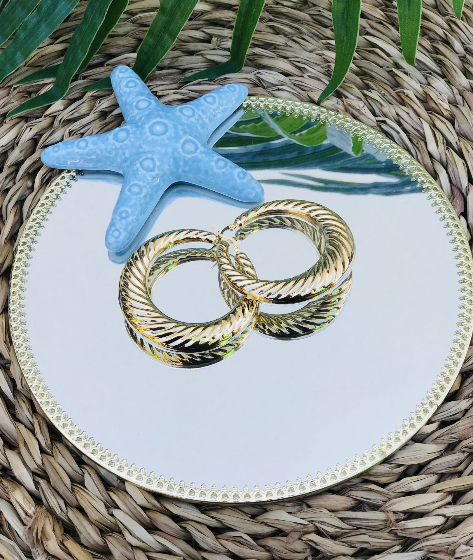 Brincos de argola dourados com textura torcional sobre espelho redondo com moldura dourada e decoração de estrela-do-mar azul.