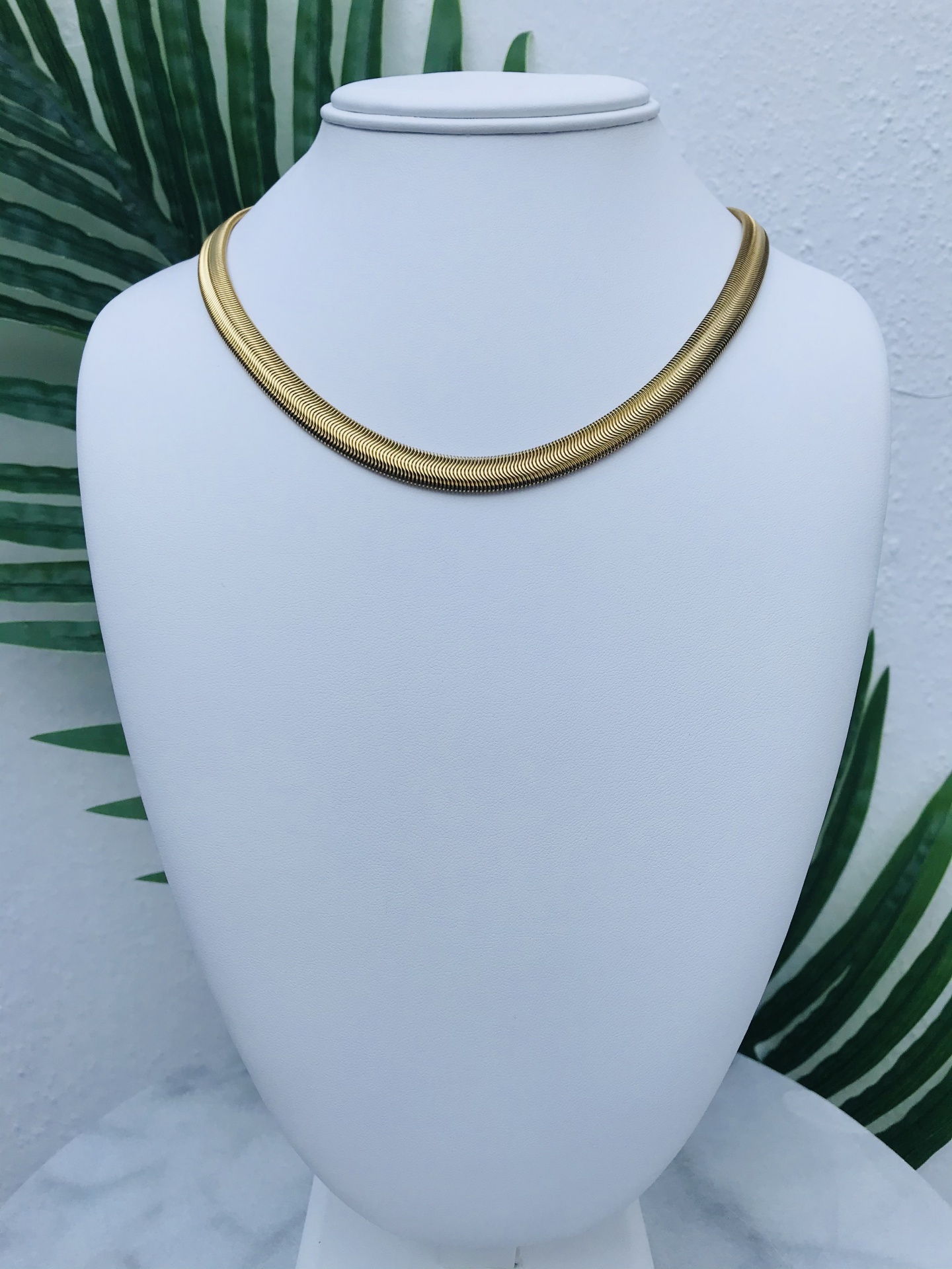 Colar dourado liso exibido em manequim branco com fundo de folhas verdes.