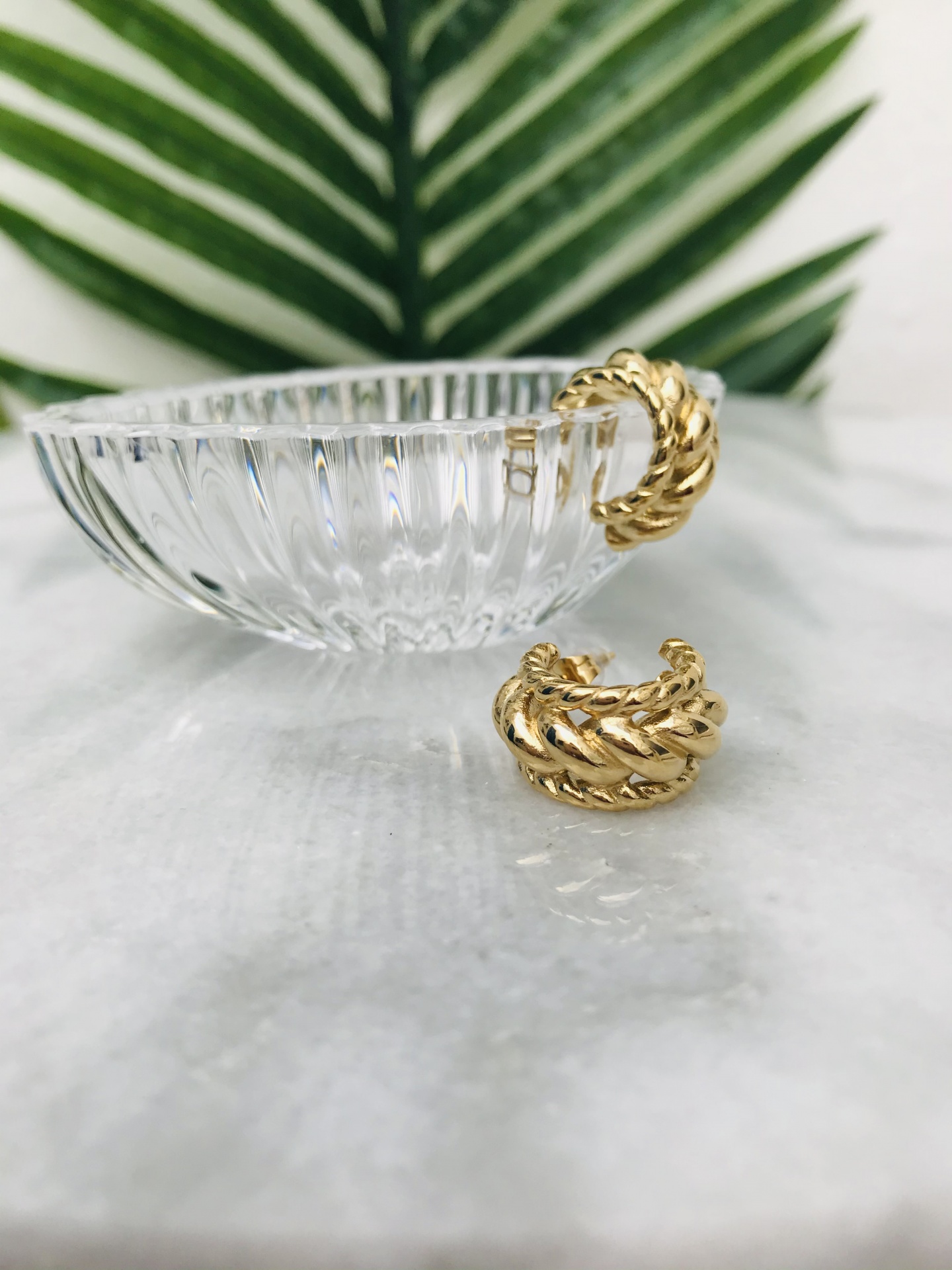 Brincos dourados trançados com taça de vidro e folha verde ao fundo