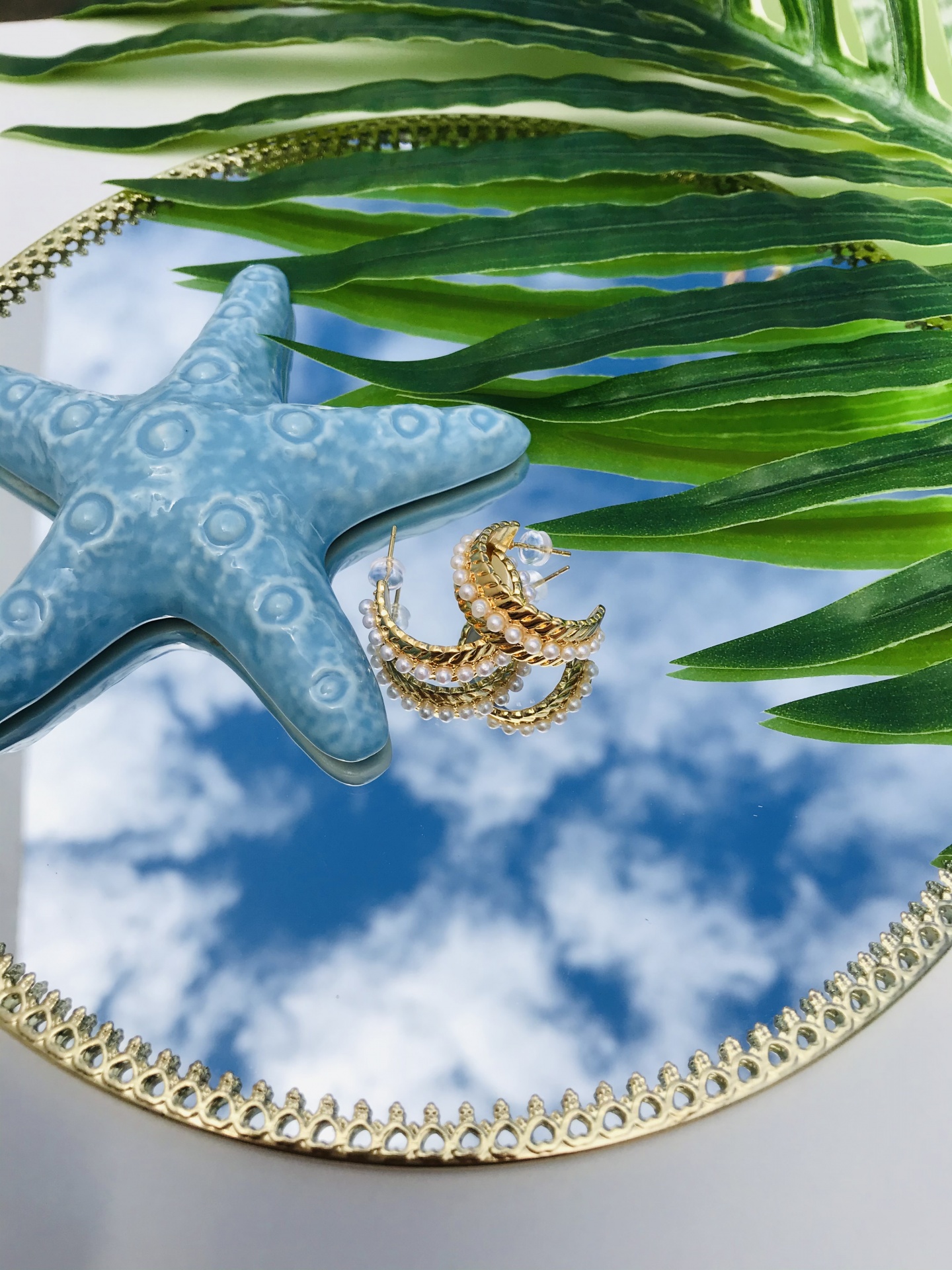 Espelho redondo dourado com reflexo do céu, estrela do mar azul e brincos dourados