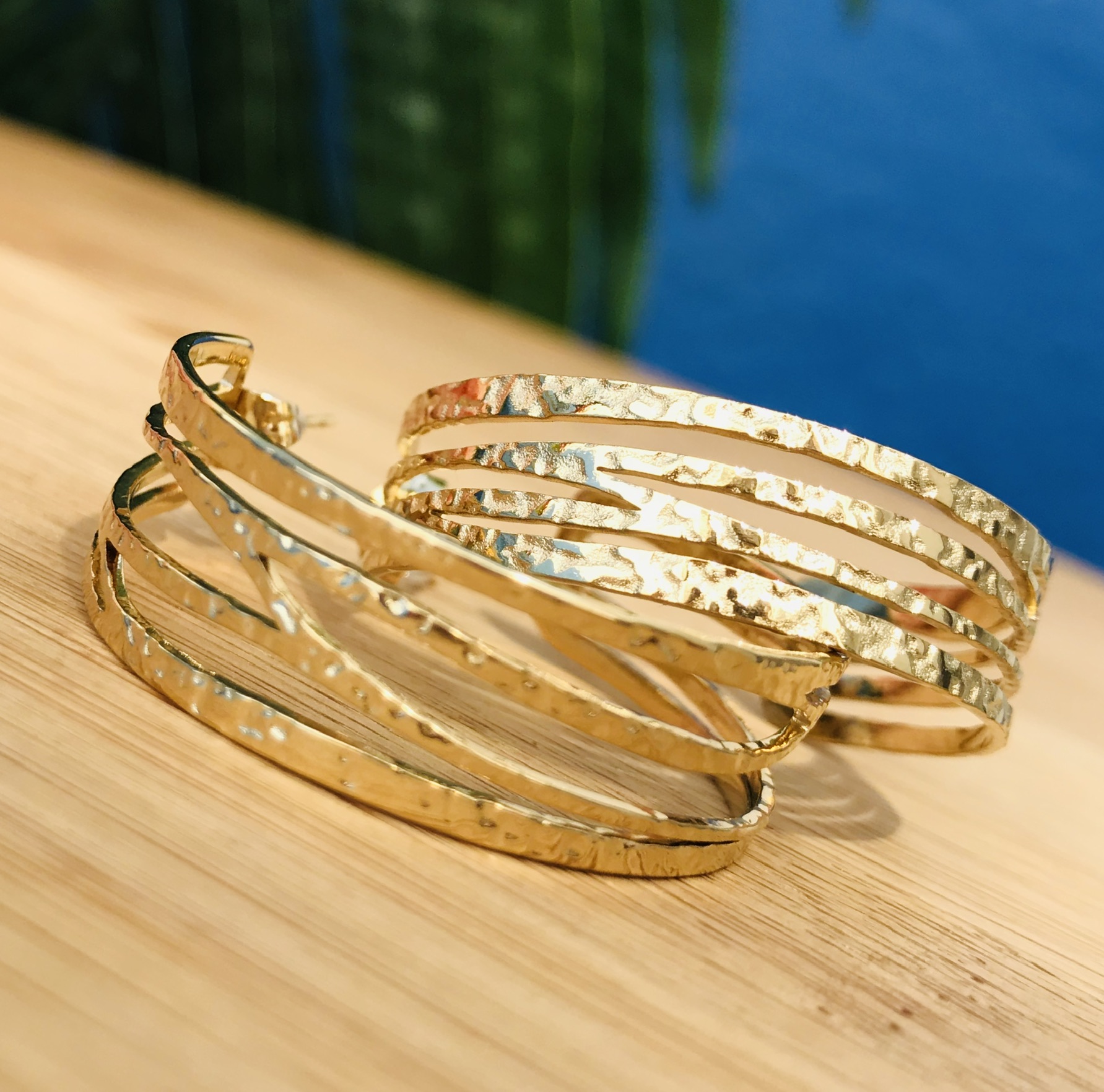 Brincos de argola dourados com textura martelada sobre madeira clara e fundo de piscina azul