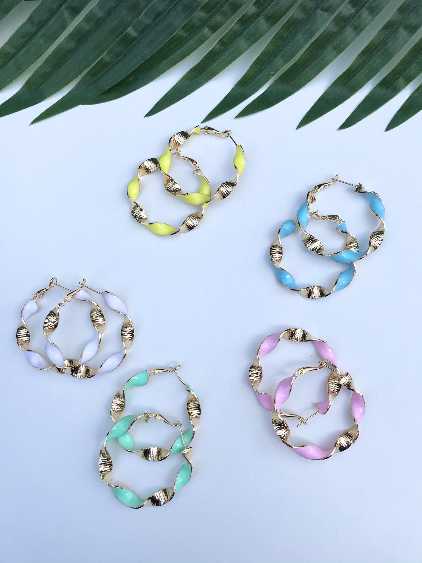 Cinco pares de brincos de argola coloridos torcidos com partes douradas sobre fundo branco e folha verde