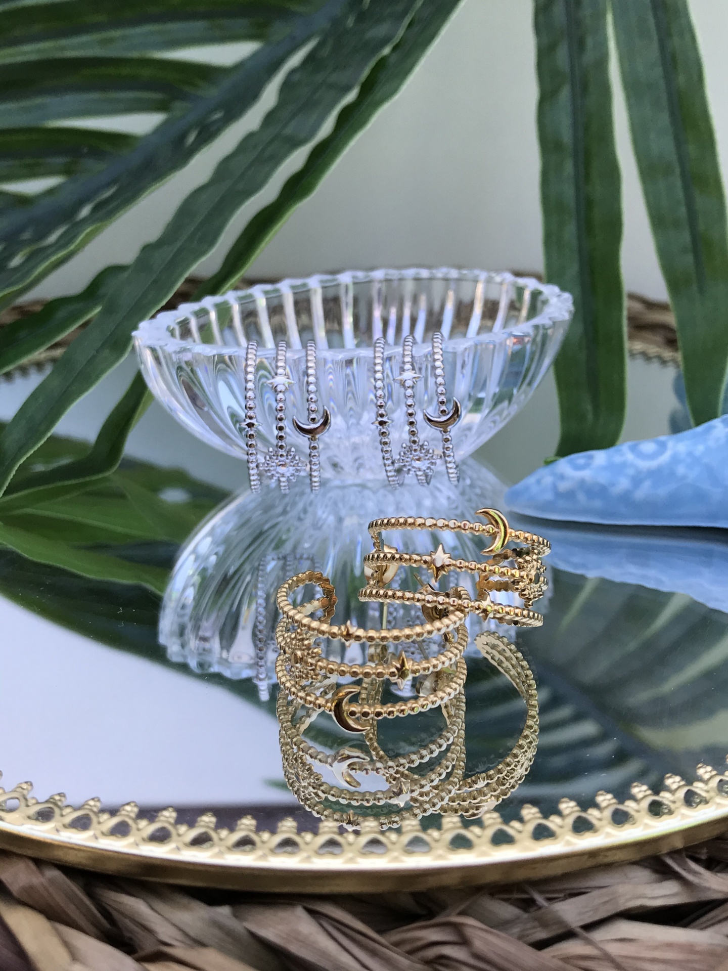 Brincos de argola dourados e prateados com lua crescente num espelho com moldura dourada e folhas verdes no fundo