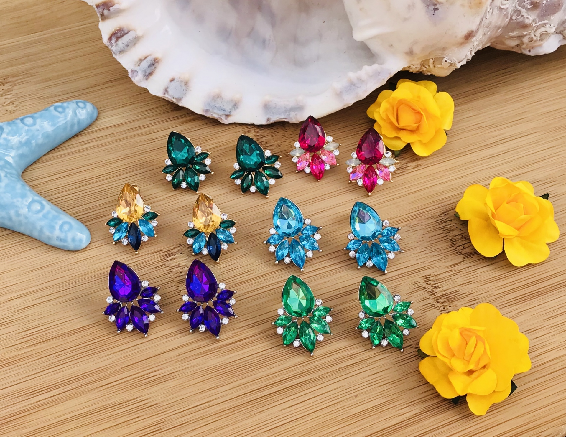 Pares de brincos coloridos em superfície de madeira com flores amarelas, concha branca e estrela do mar azul