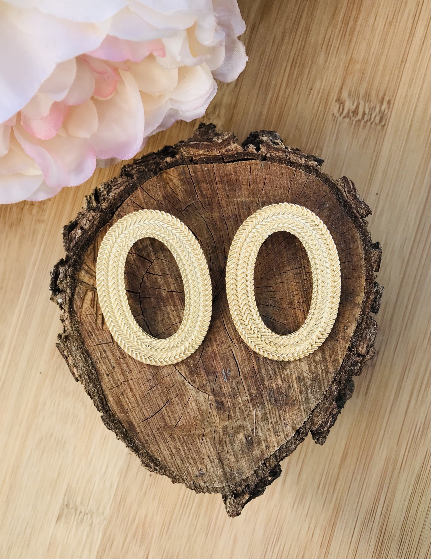 Brincos ovais dourados com textura trançada sobre madeira rústica ao lado de flor branca e rosa