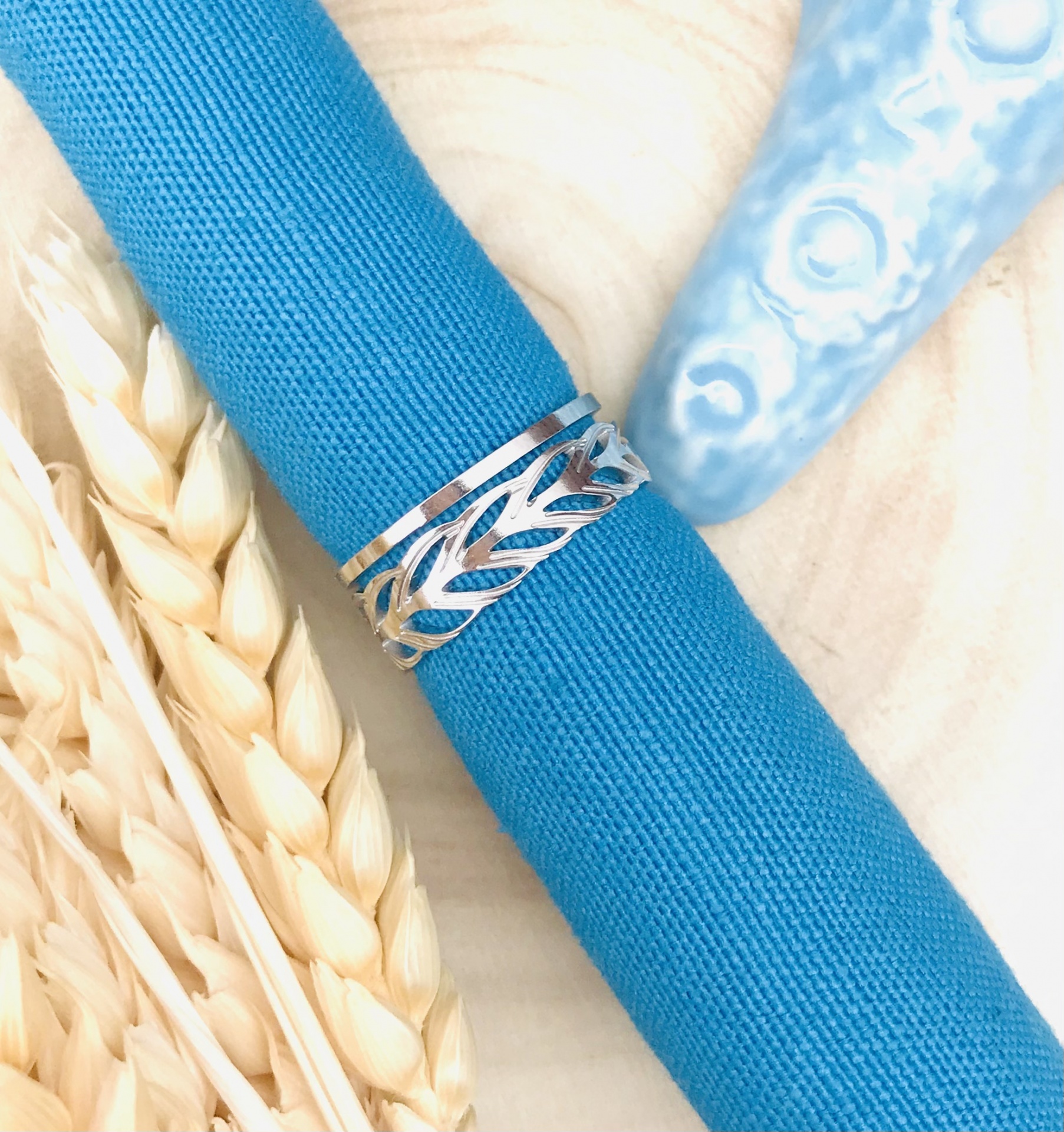 Anel prateado com design de folhas em tecido azul, espigas de trigo e concha azul clara ao fundo