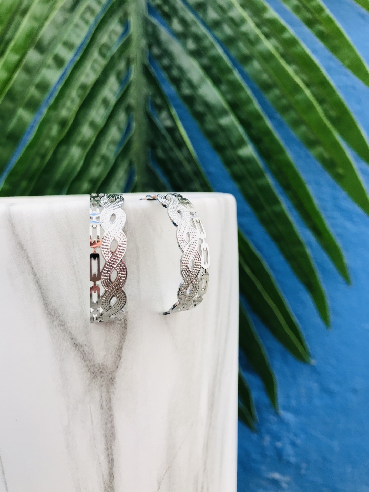 Brincos de prata entrelaçados sobre base de mármore com folhas verdes ao fundo