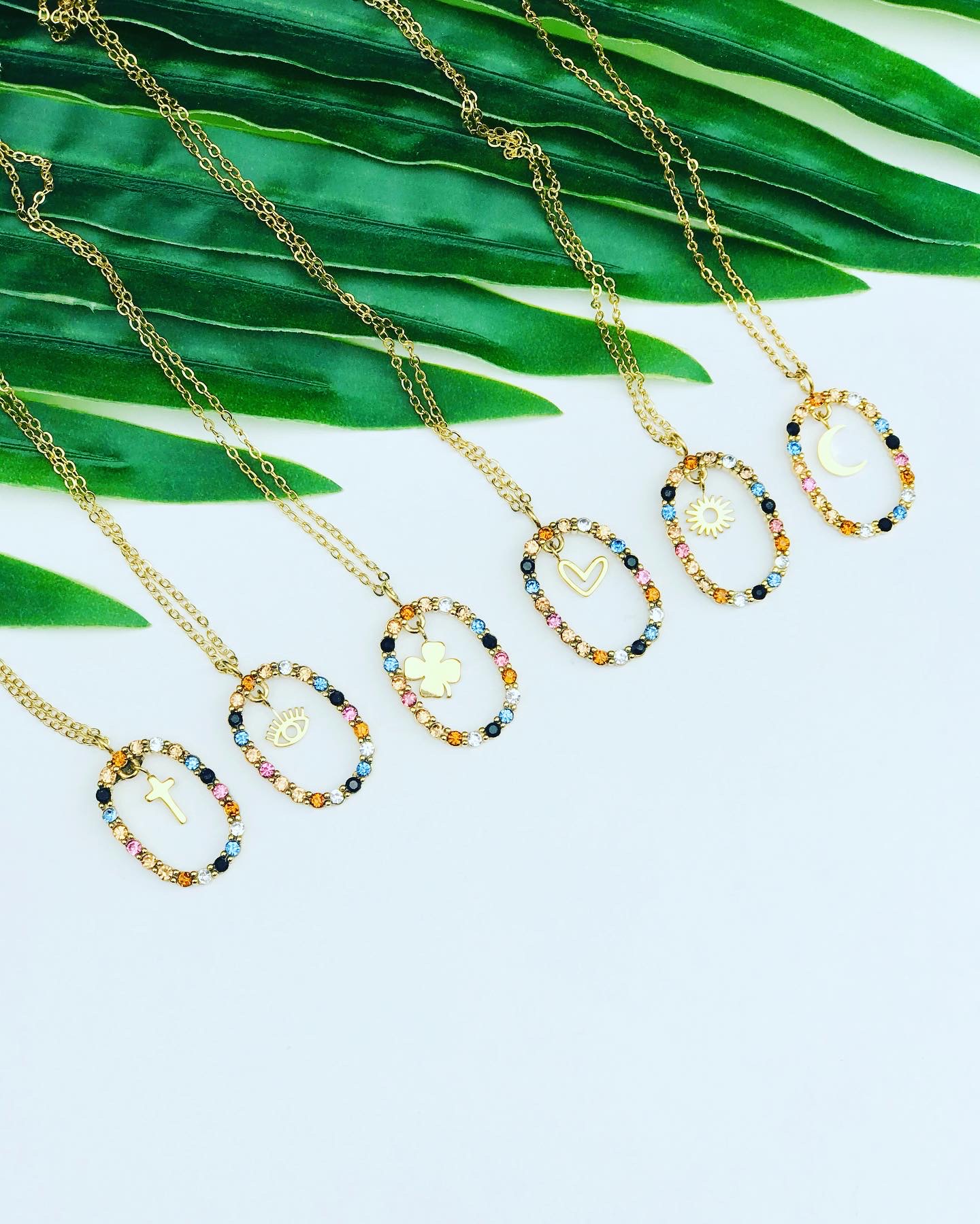 Seis colares dourados com pendentes ovais e pedras coloridas, cada um com diferente símbolo, sobre fundo branco com folhas verdes.