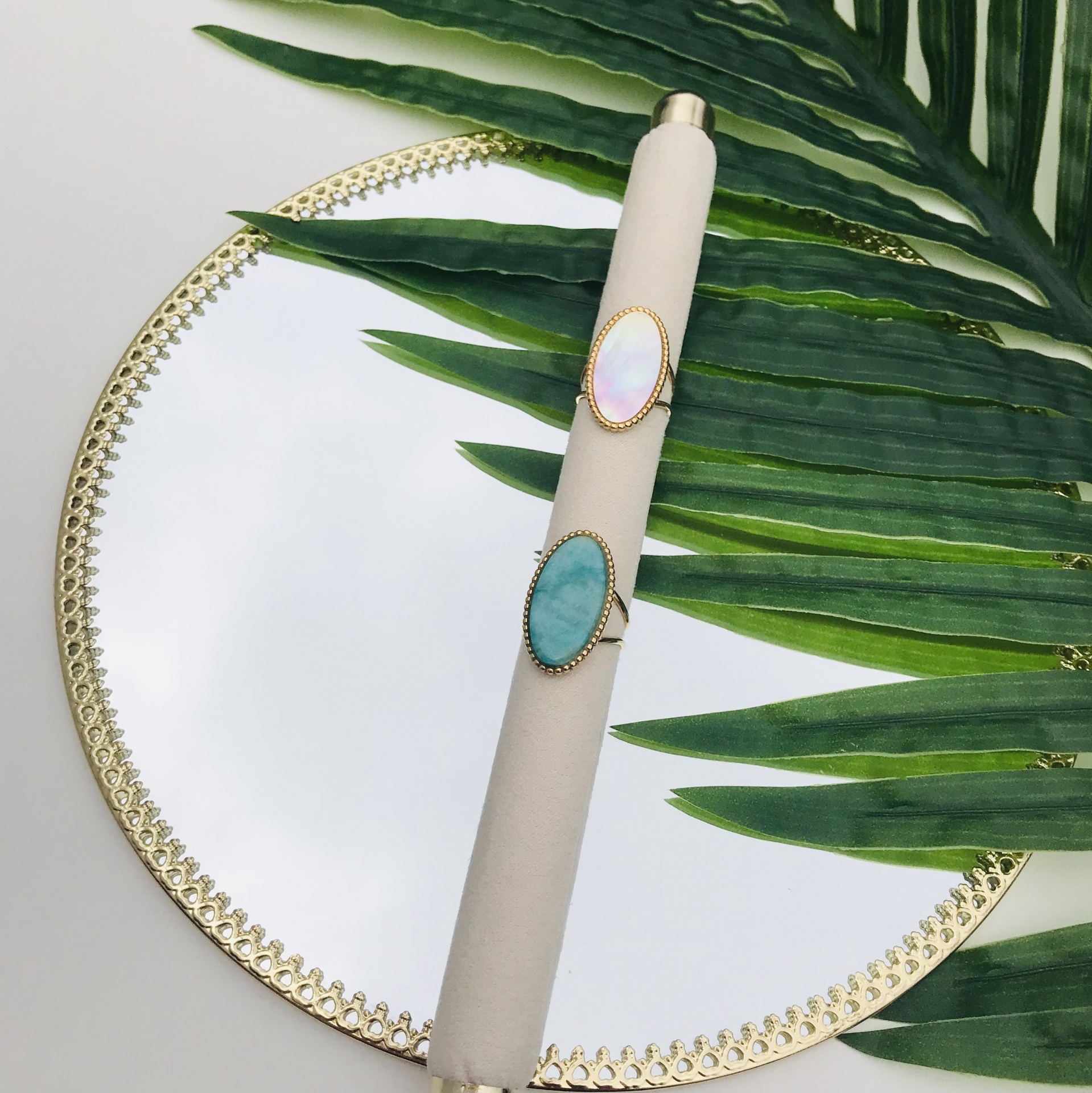 Dois anéis com pedras ovais em suporte creme sobre espelho redondo com borda dourada e folha verde