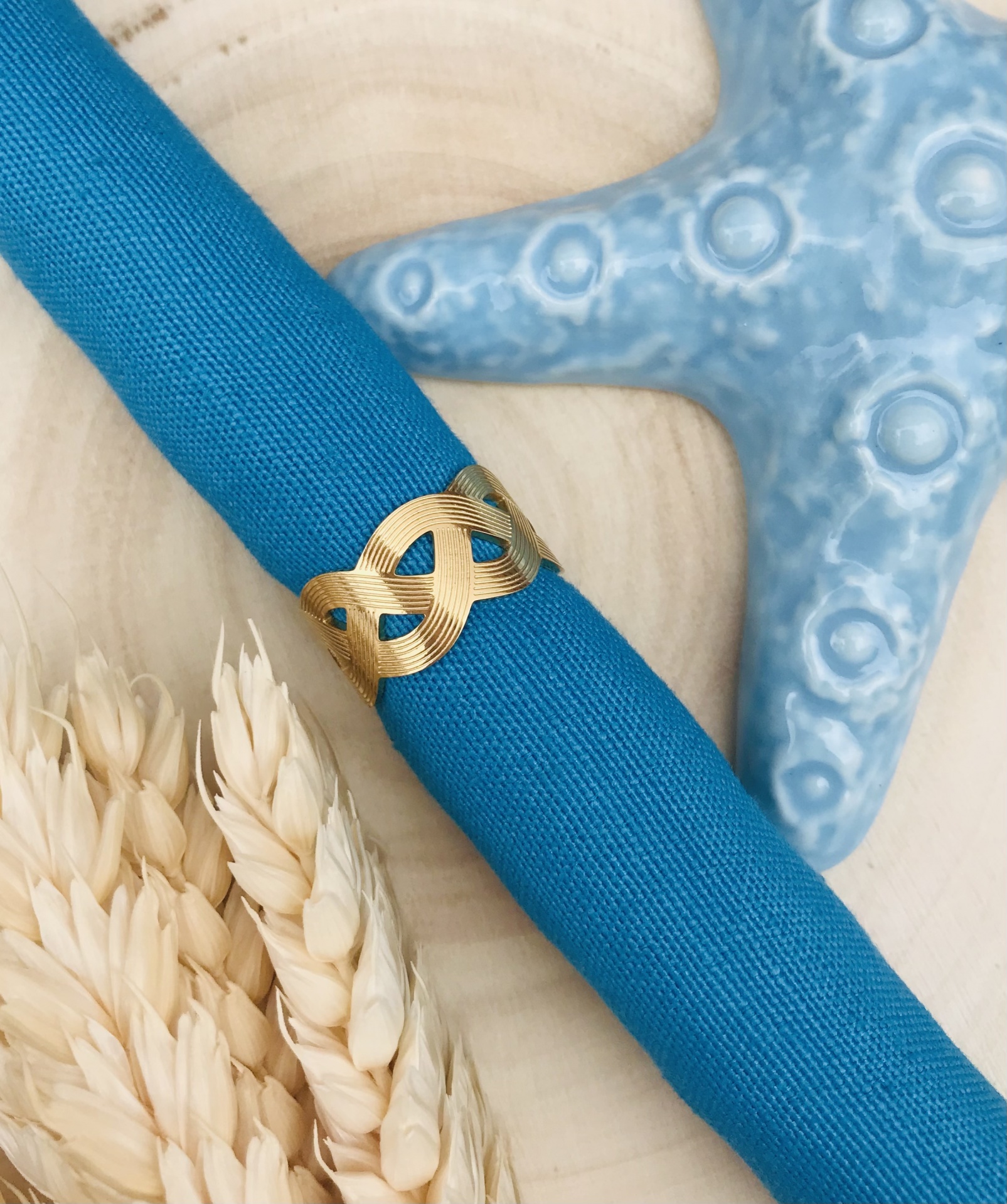 Guardanapo azul com anel dourado, estrela do mar azul e espigas claras sobre madeira clara