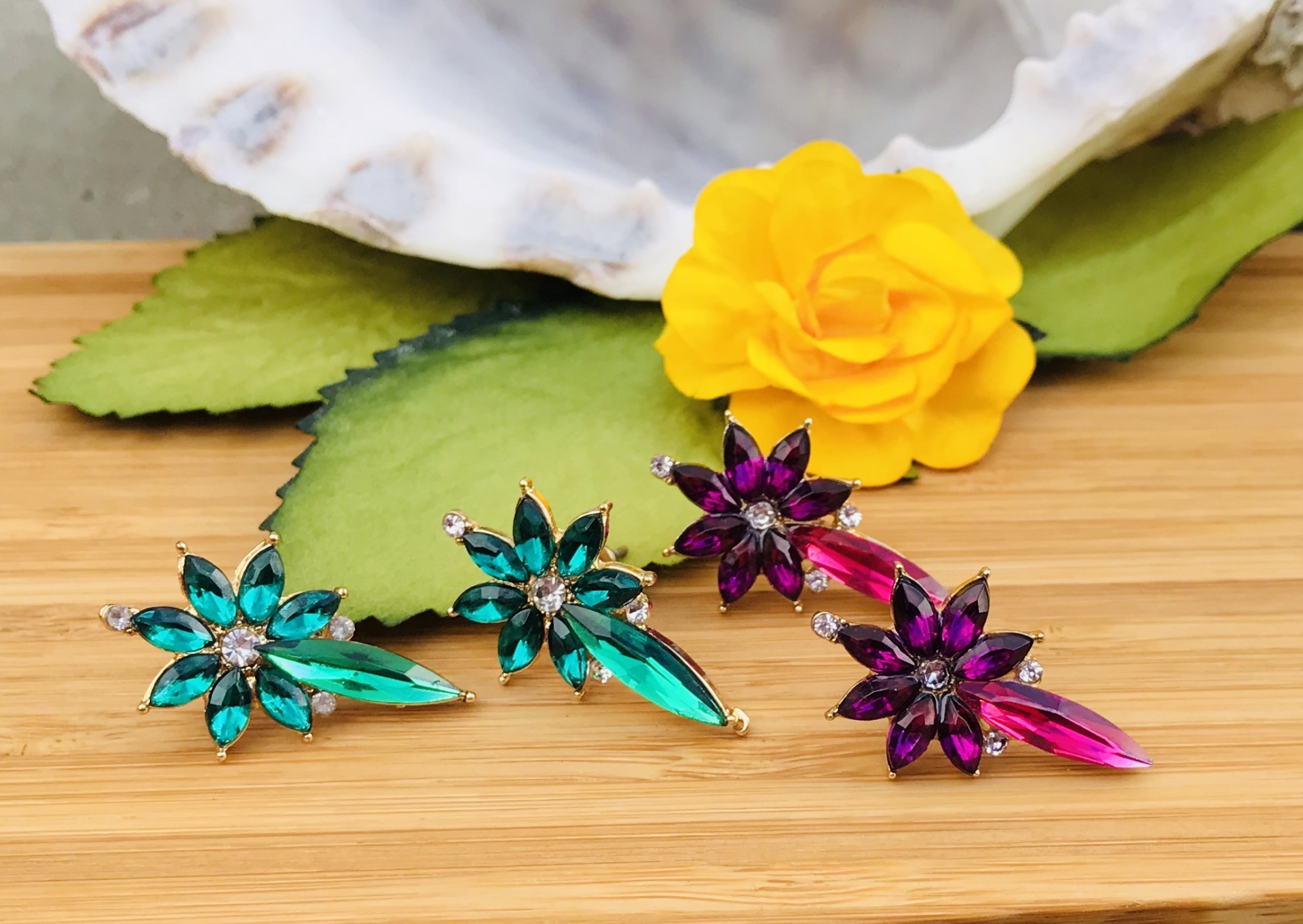 Brincos florais verde e roxo sobre madeira com flor amarela e folha verde ao fundo