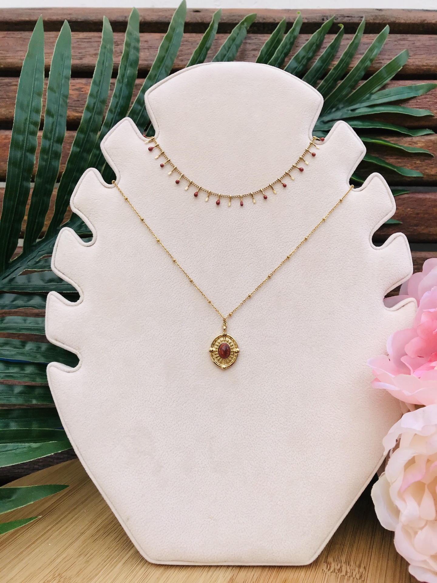 Dois colares dourados num suporte branco com folhas verdes e flores cor de rosa ao fundo