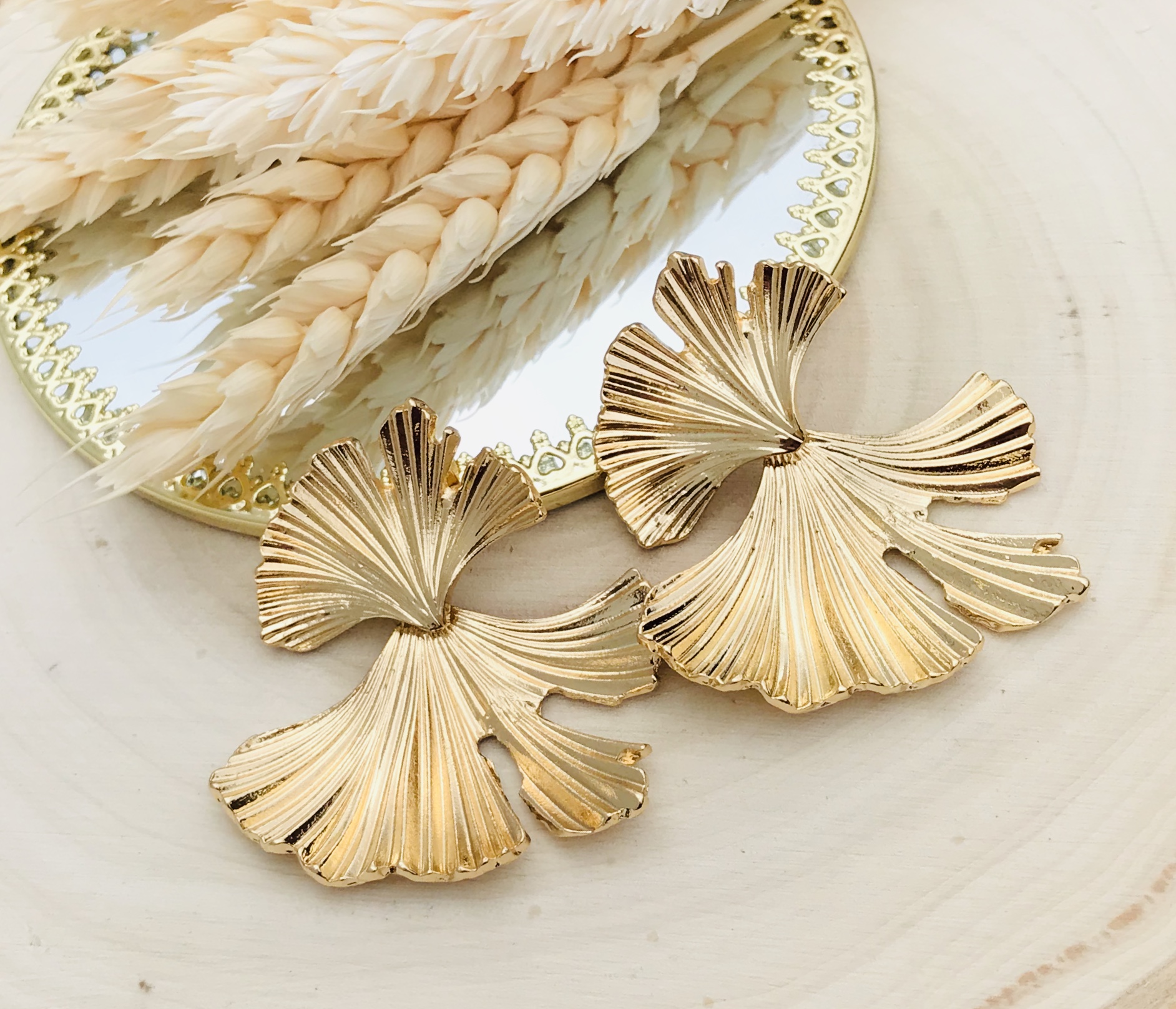 Brincos dourados em forma de folhas sobre madeira clara junto a espelho e ramos secos