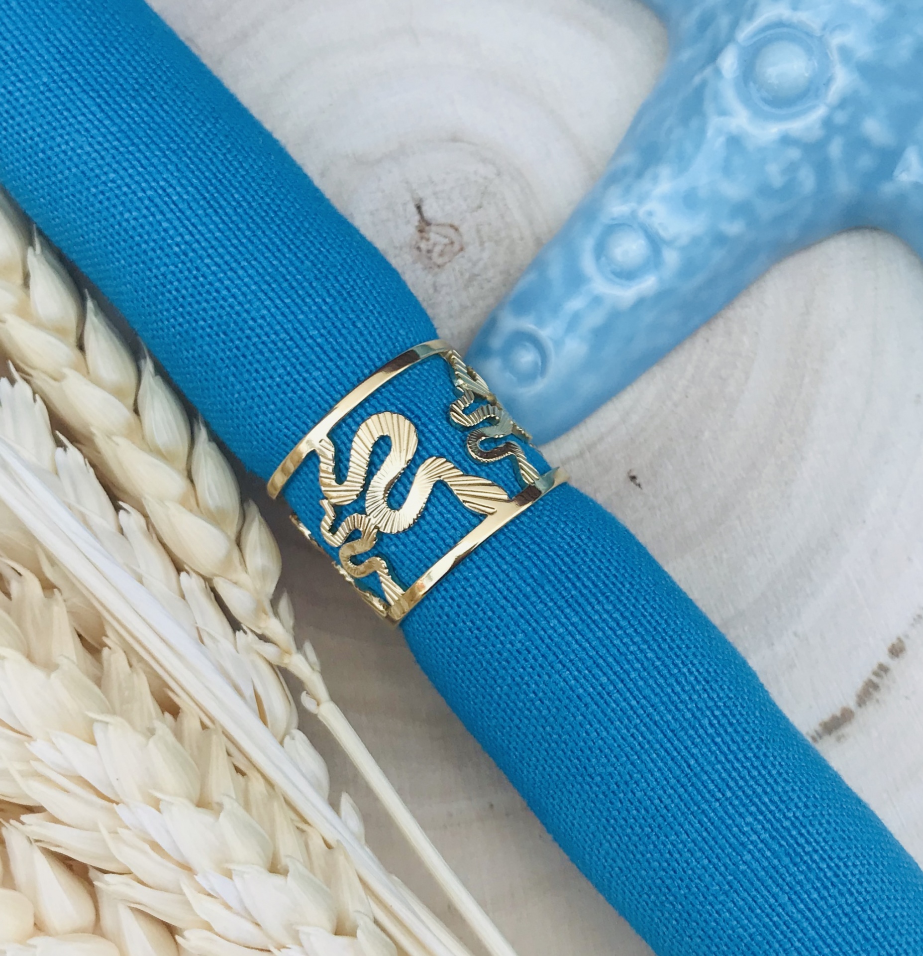 Anel dourado de cobra enrolada em tecido azul com fundo de madeira clara e elementos naturais