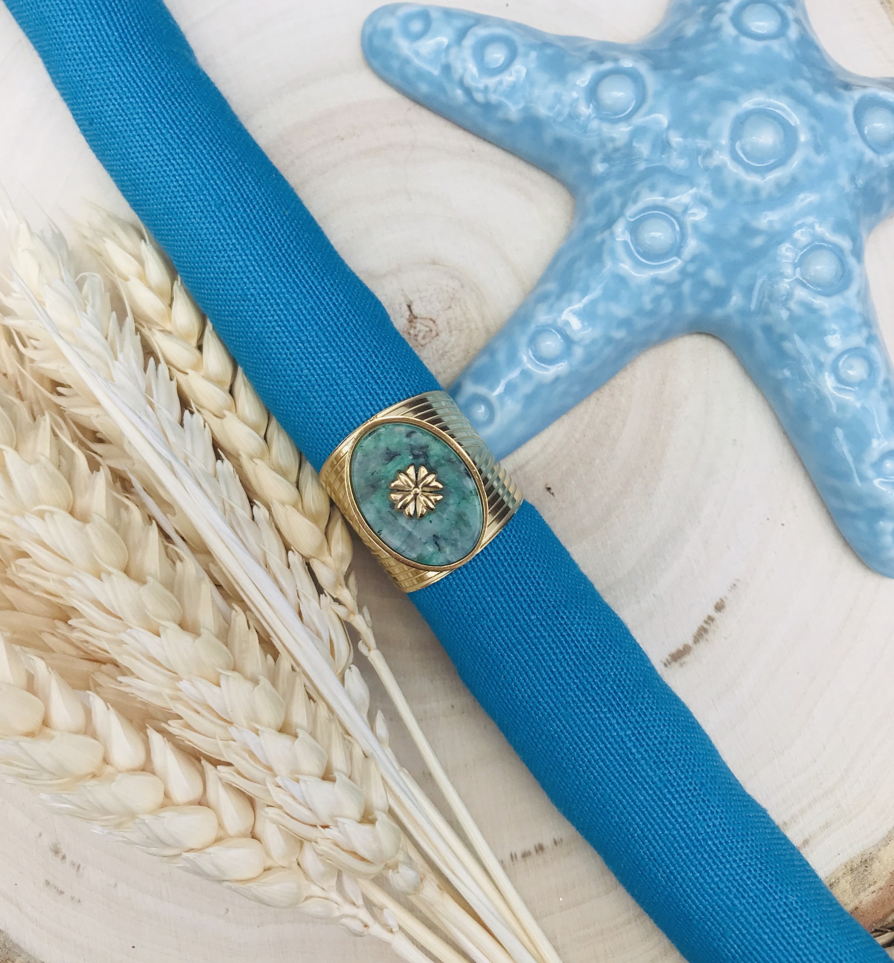 Guardanapo azul com anel decorativo dourado e pedra verde, espigas de trigo e estrela-do-mar azul sobre madeira clara