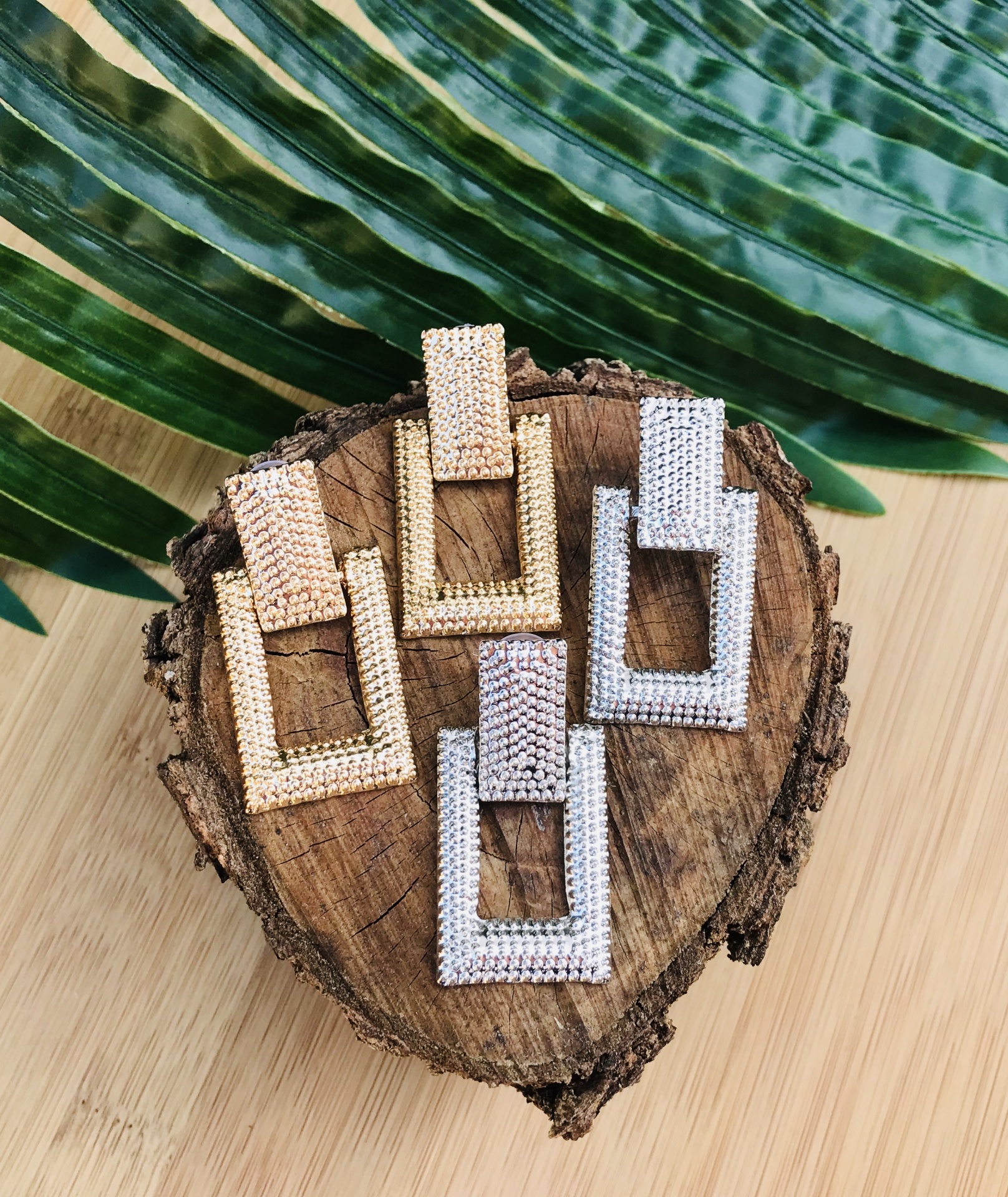 Dois pares de brincos retangulares dourados e prateados sobre madeira com folhas verdes ao fundo