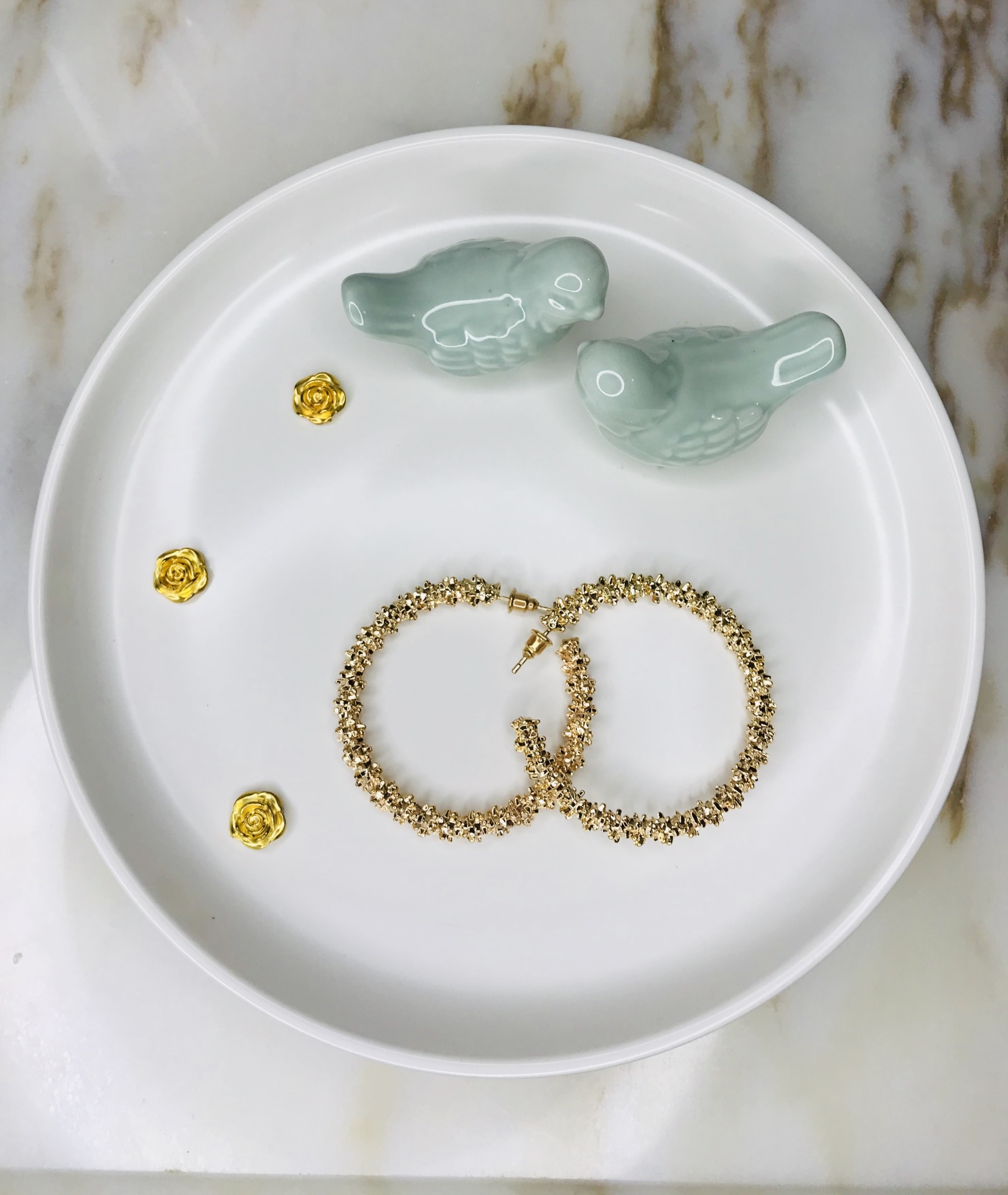 Brincos dourados, pendentes florais dourados e figuras de pássaros verdes num prato branco