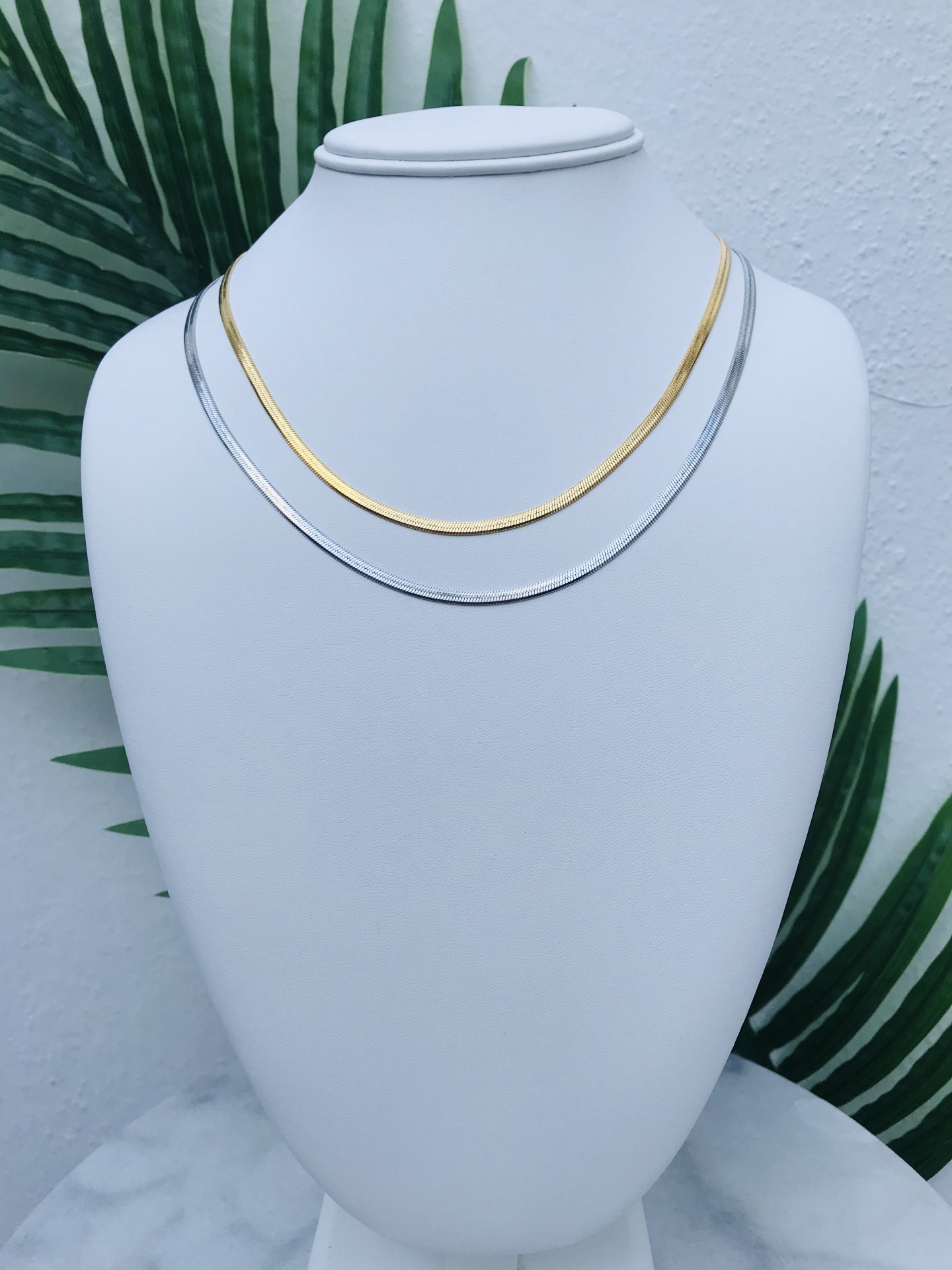 Colares dourado e prateado em suporte branco com fundo de folhas verdes