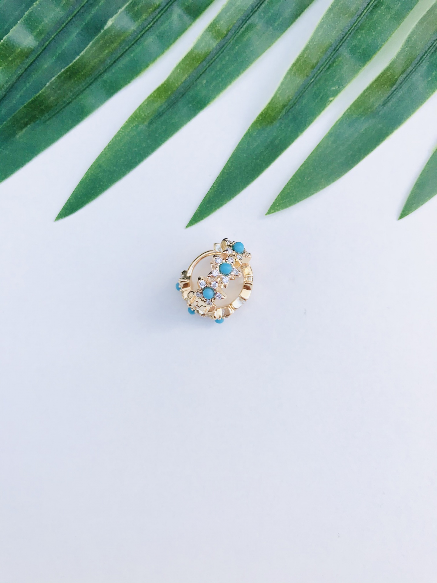 Brinco dourado com pedras azuis e brancas sobre fundo branco e folhas verdes