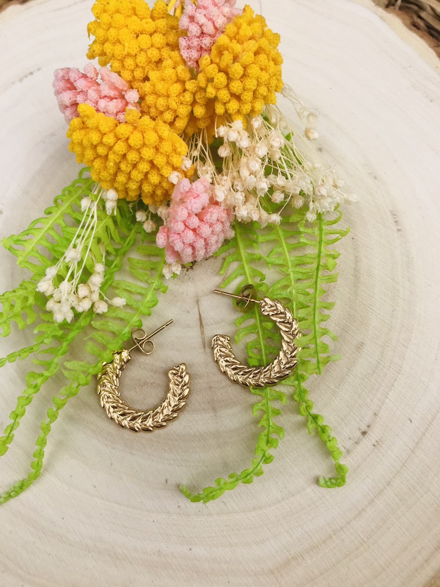 brincos dourados em argola com textura trançada em fundo de madeira com flores artificiais