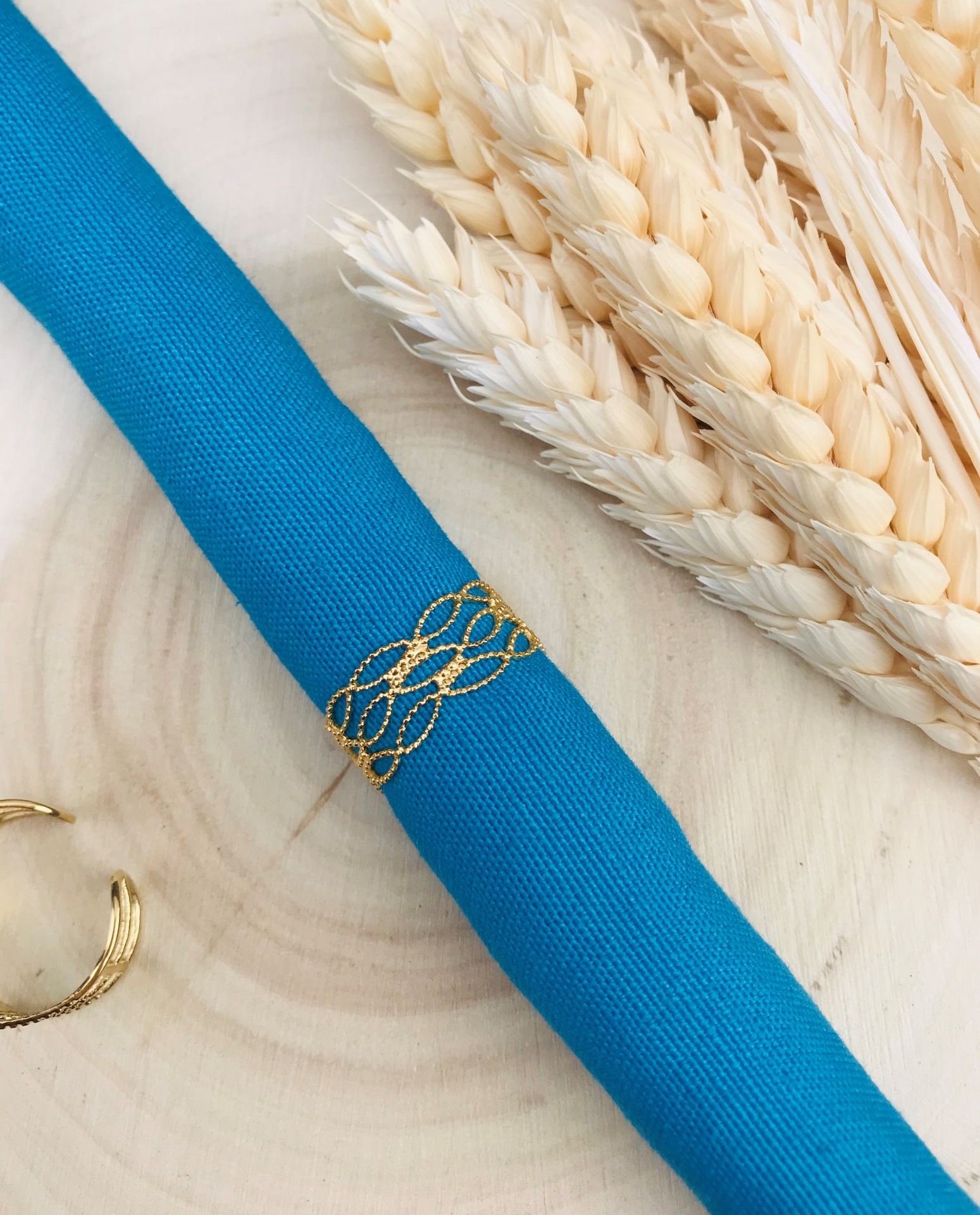 Anel dourado entrelaçado sobre tecido azul com espigas claras num fundo de madeira