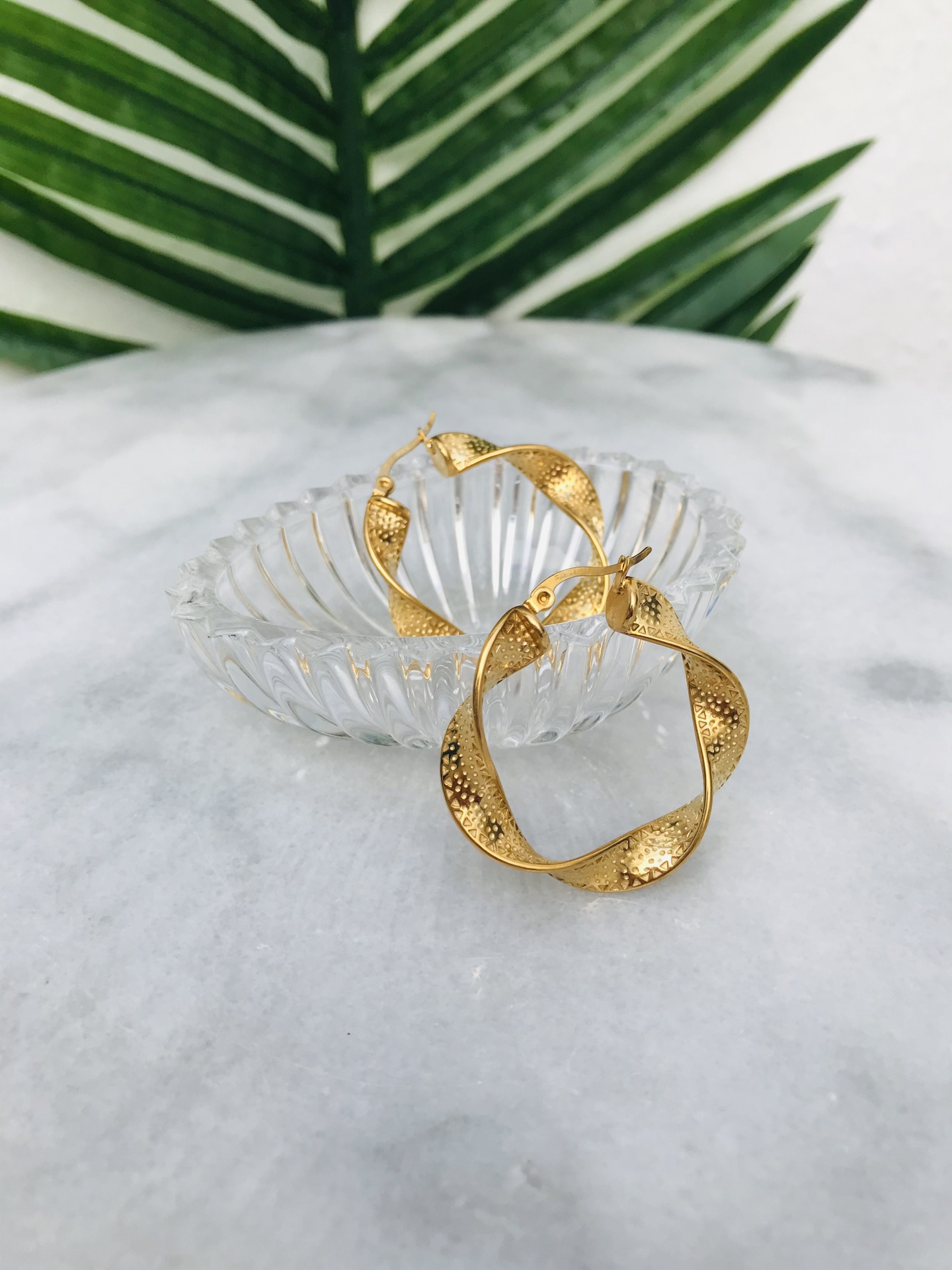 Brincos de ouro texturizados em prato de vidro sobre mármore branco com folhas verdes ao fundo