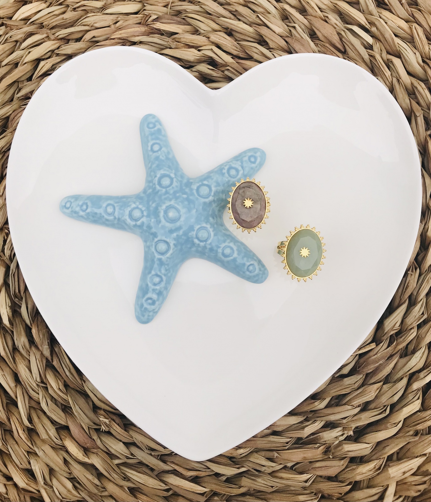 Prato branco em forma de coração com estrela-do-mar azul e dois anéis coloridos sobre tapete de palha