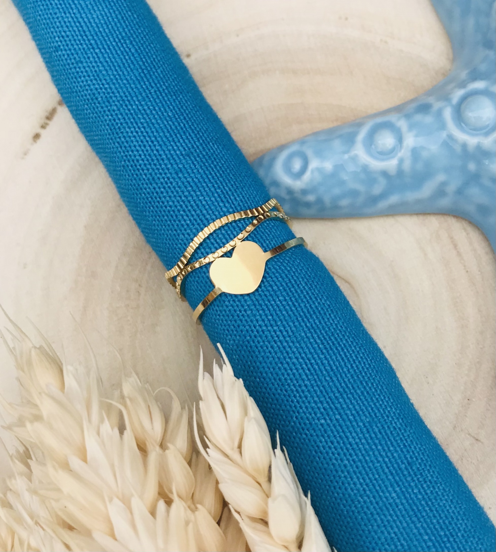 Anel dourado com coração em pano azul sobre base de madeira com trigo seco e estrela do mar