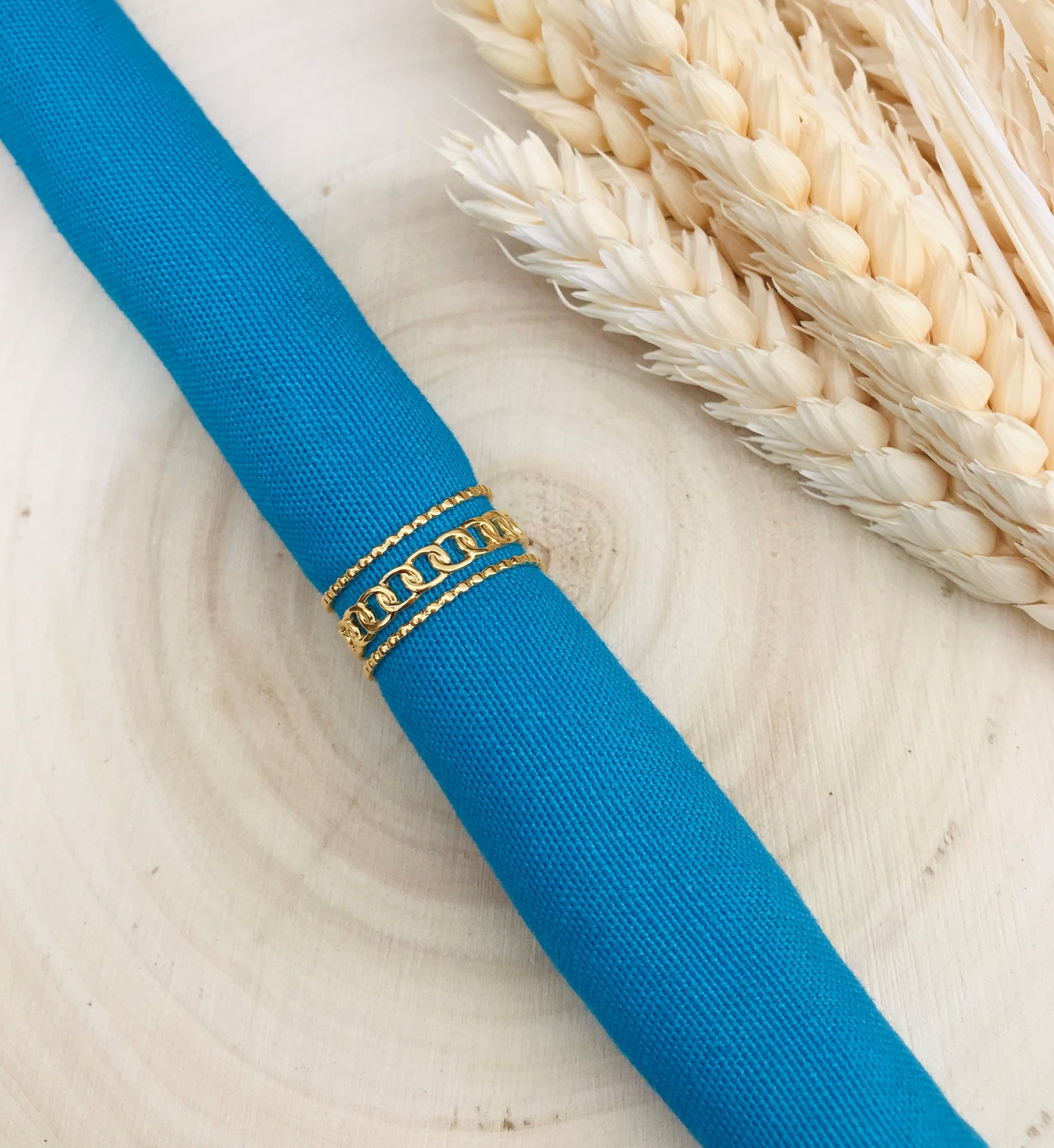 Argola dourada com corrente num guardanapo azul junto a espigas de trigo