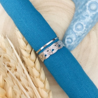 Anel prateado entrelaçado com pedras num guardanapo azul sobre madeira clara com espigas de trigo e objeto azul