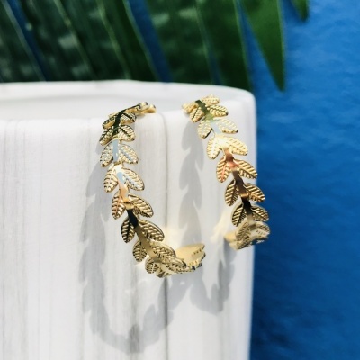 Brincos dourados em padrão de folhas numa superfície branca com fundo azul e folhas verdes.