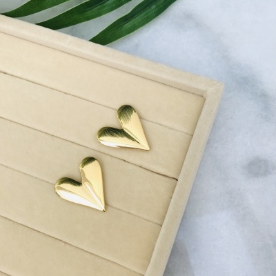 Brincos de coração dourados sobre base de veludo bege com folhas verdes ao fundo