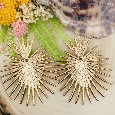 Brincos dourados com design de folhas secas, sobre madeira com conchas e flores artificiais.