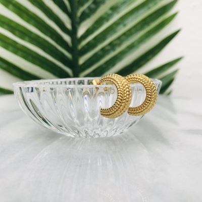 Brincos dourados em forma de meia-lua sobre tigela de vidro com folha verde ao fundo.