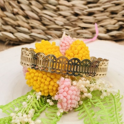 Pulseira dourada com padrão recortado sobre flores decorativas amarelas e cor-de-rosa com folhas, em fundo de madeira e tapete de palha.