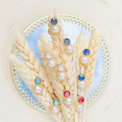 Brincos dourados com pérolas e pedras coloridas sobre espigas de trigo secas em espelho.