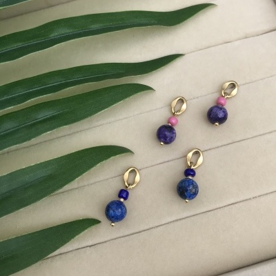 Dois pares de brincos com contas coloridas e fecho dourado sobre fundo de tecido bege e folhas verdes