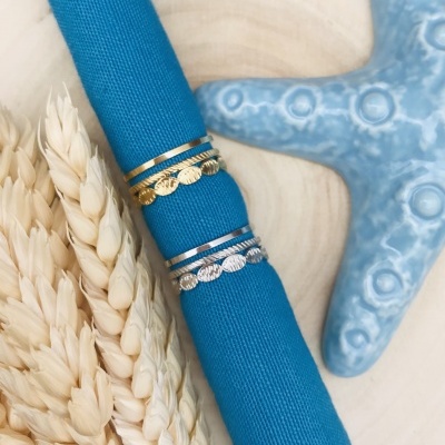 Anéis dourados e prateados com discos texturizados em tecido azul, estrela-do-mar azul e espigas de trigo secas sobre madeira.