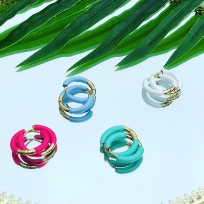 Quatro pares de brincos coloridos em argola com detalhes dourados sobre um espelho redondo com borda dourada, com folhas verdes ao fundo.