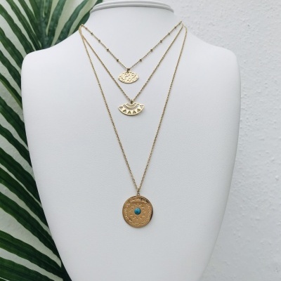 Colar triplo dourado com três pendentes em busto branco e folhas verdes ao fundo