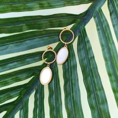 Brincos dourados com pendentes ovais brancos sobre folha verde