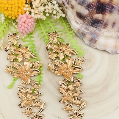 Brincos dourados longos com flores em fundo com plantas, flores secas e concha