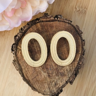 Brincos ovais dourados com textura trançada sobre madeira rústica ao lado de flor branca e rosa