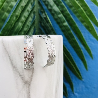 Brincos de prata entrelaçados sobre base de mármore com folhas verdes ao fundo