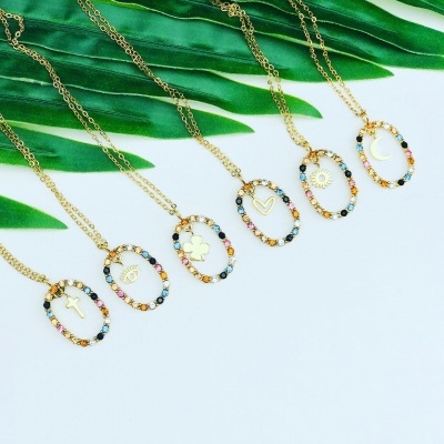 Seis colares dourados com pendentes ovais e pedras coloridas, cada um com diferente símbolo, sobre fundo branco com folhas verdes.